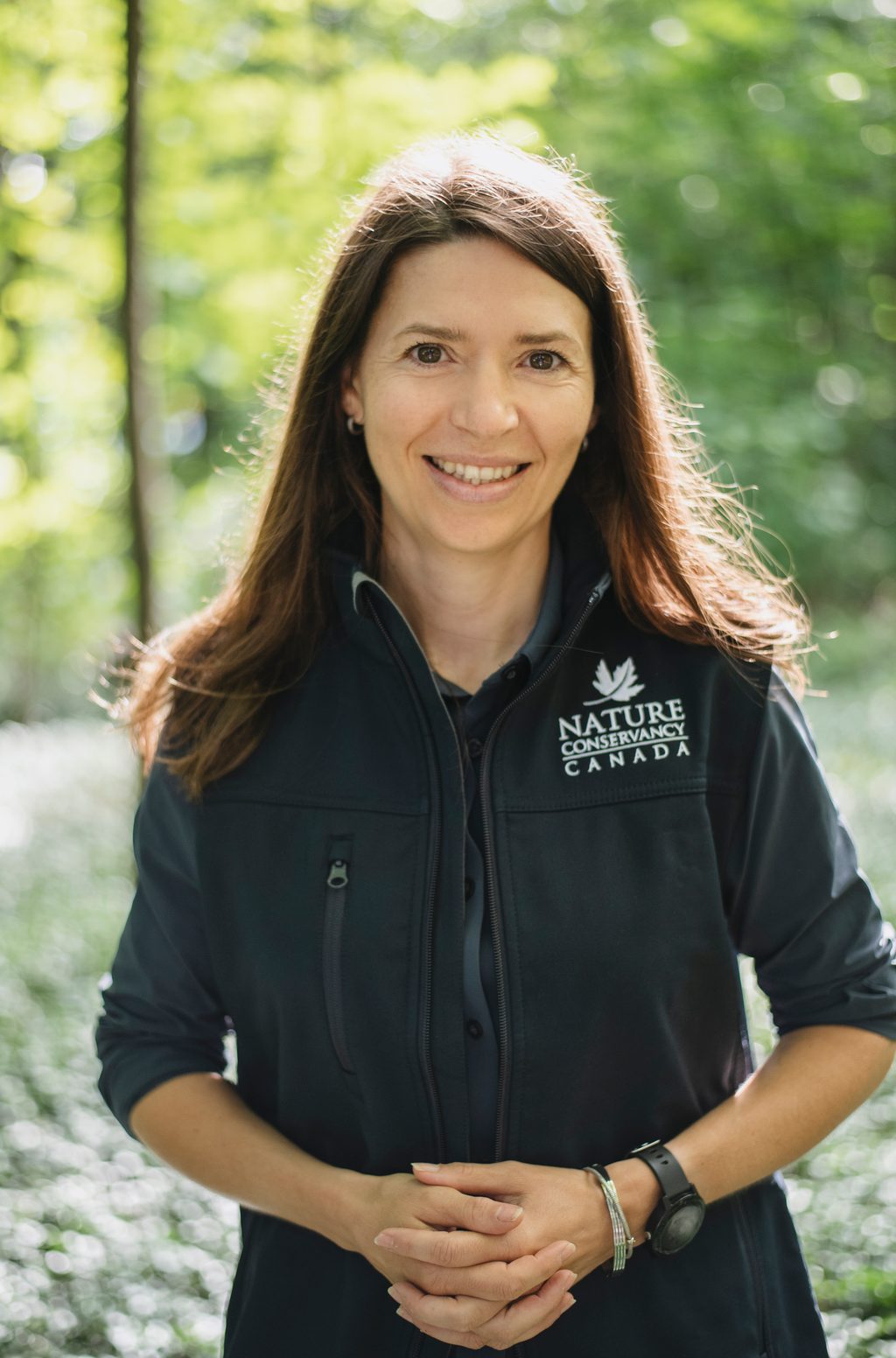 A woman wearing a Nature Conservancy Canada jacket smiles in a wooded area.