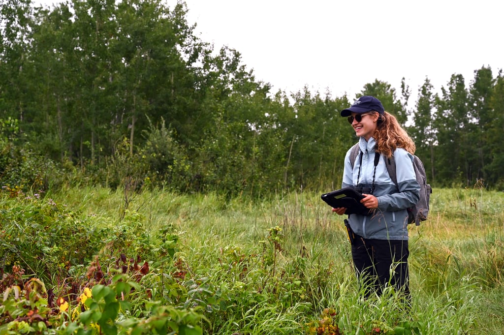 A woman with a backpack and clipboard stands in a grassy field near a forest.
