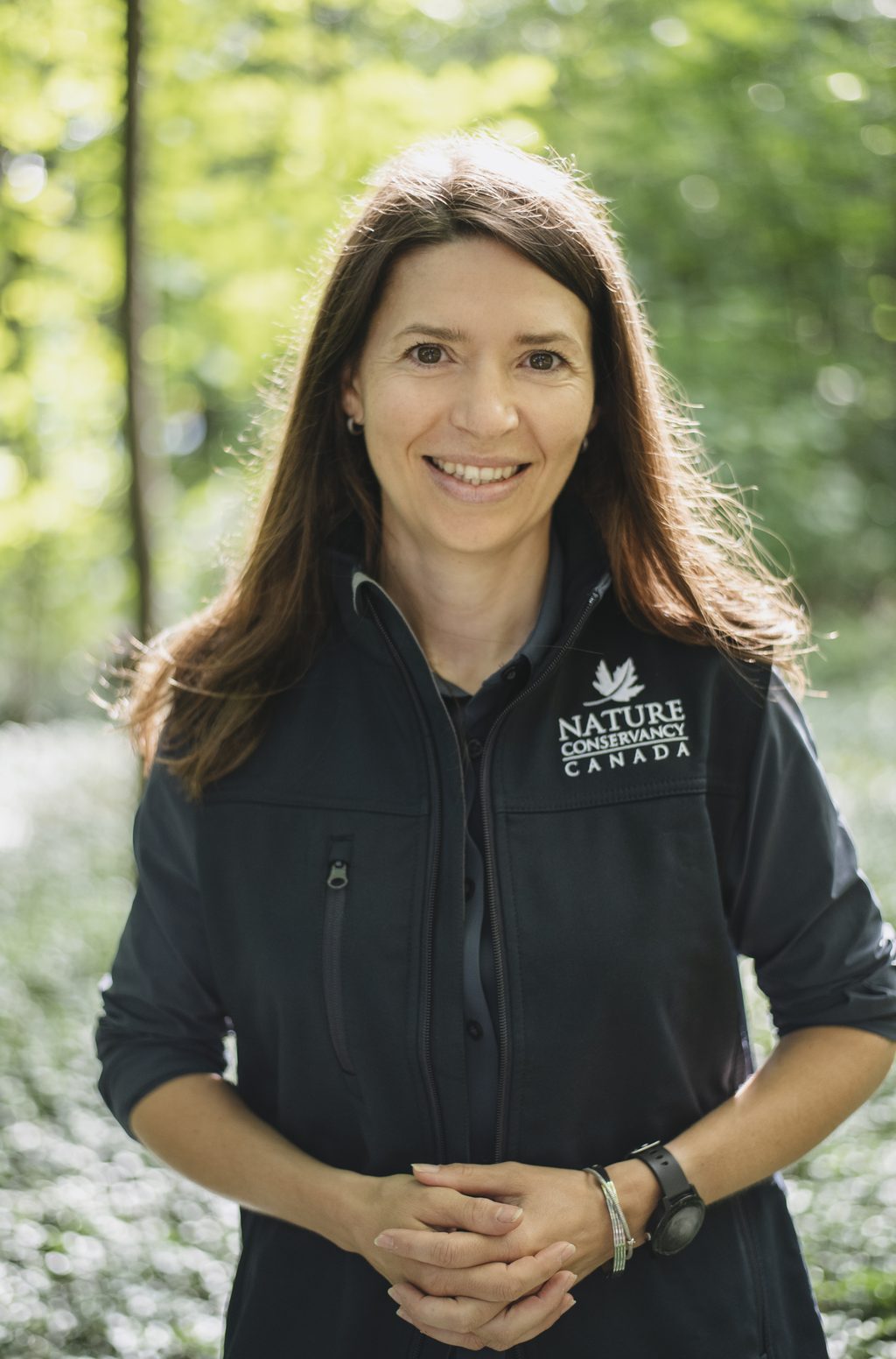 A woman wearing a Nature Conservancy Canada jacket smiles in a wooded area.