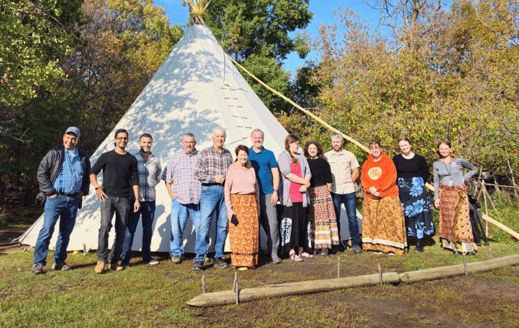 A group of people posing for a photo in front of a large white tipi in a wooded area.