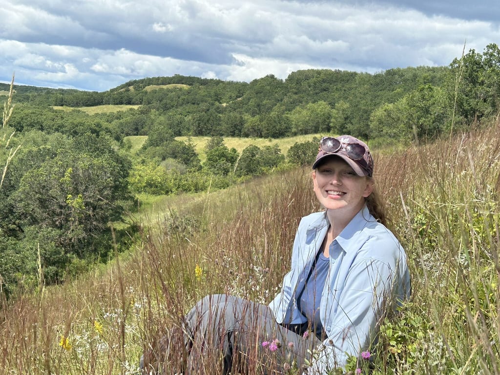 A woman sits in a grassy meadow with rolling green hills and forests in the background.