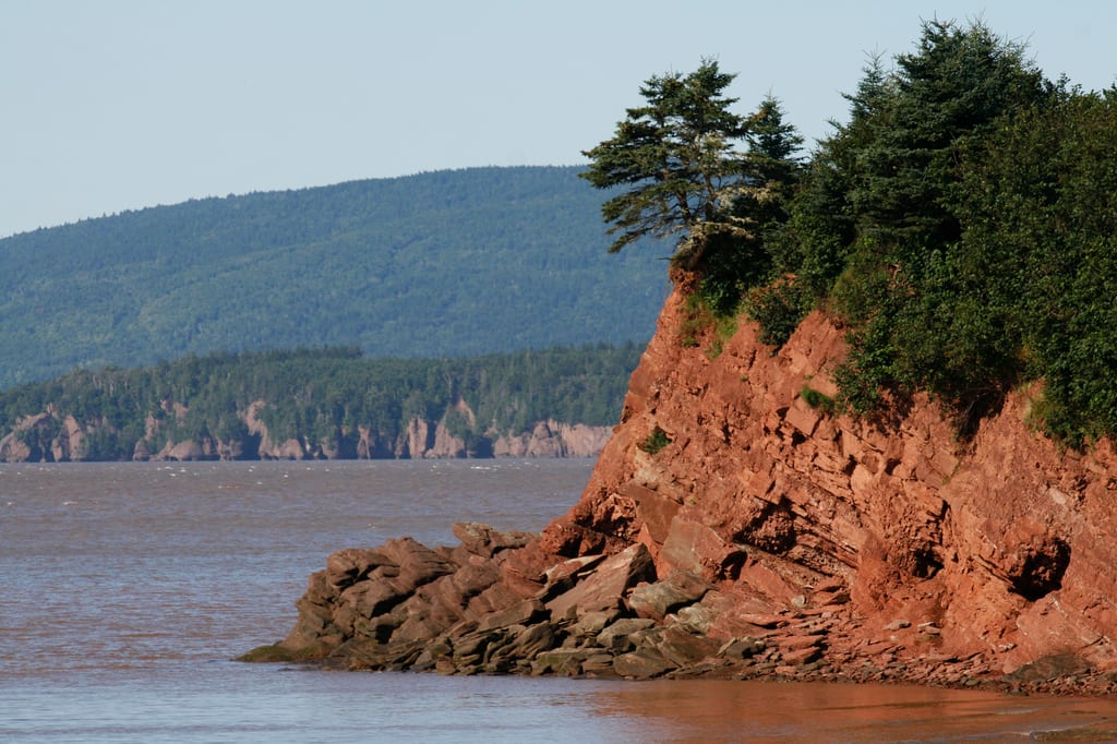 A red rocky cliff with green trees overlooks a body of water with forested hills in the distance.