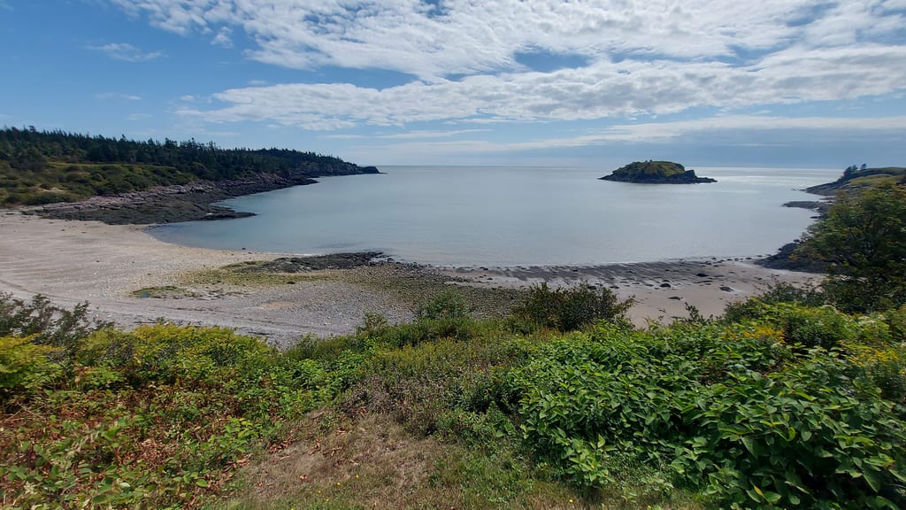 A calm bay with a sandy beach, forested hills, and a small island under a cloudy sky.