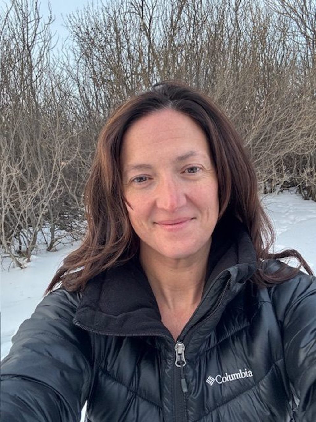 A woman in a black jacket smiles in a snowy landscape with bare bushes.