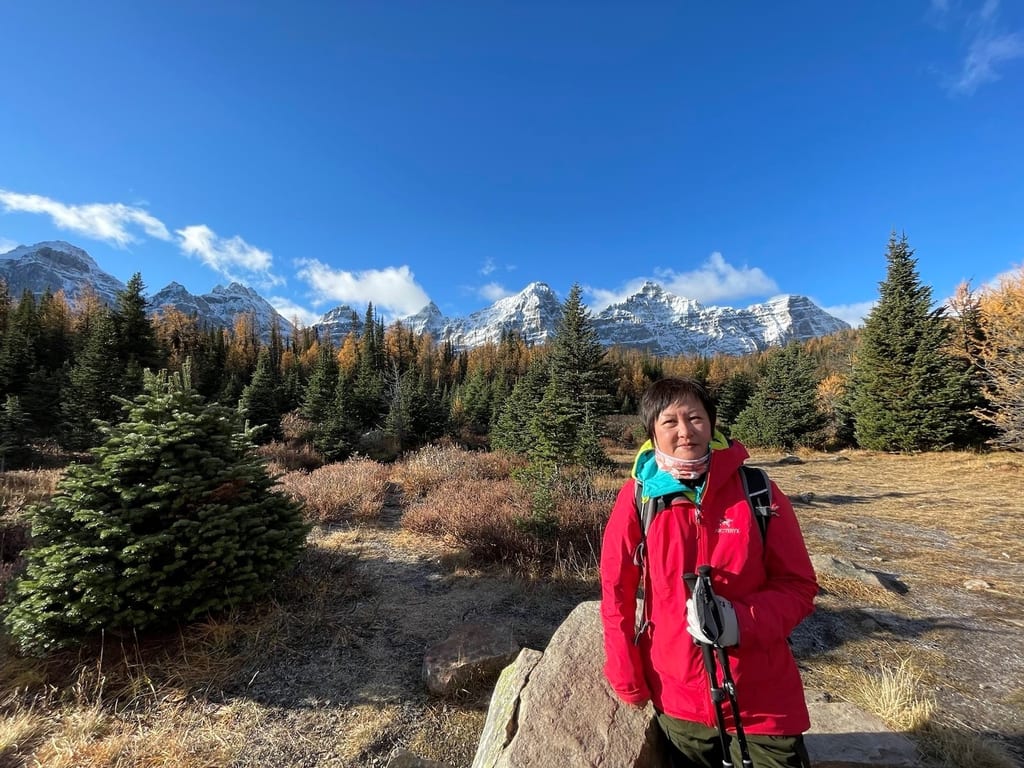 A woman in a red jacket stands in a mountainous landscape with snow-capped peaks and coniferous trees.