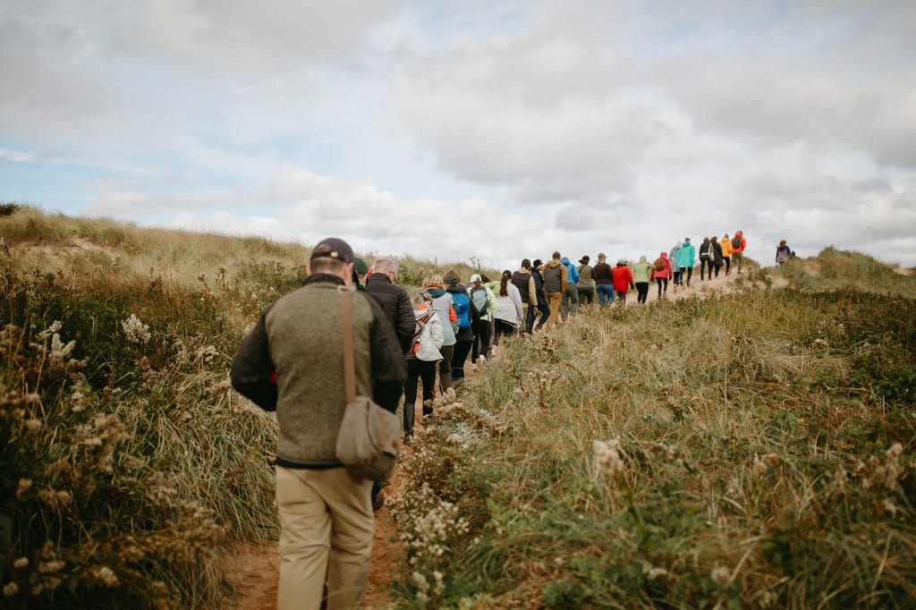 NCC staff members walk along a dirt path through grassy dunes under a cloudy sky.