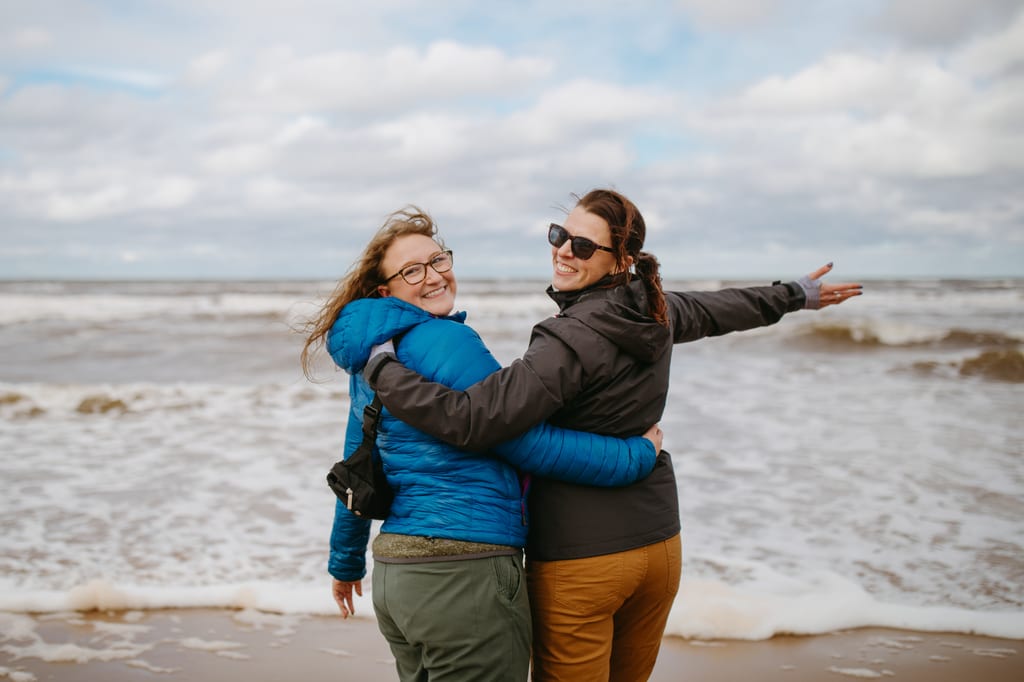 Two NCC staff members embrace and smile on a sandy beach with waves under a cloudy sky.
