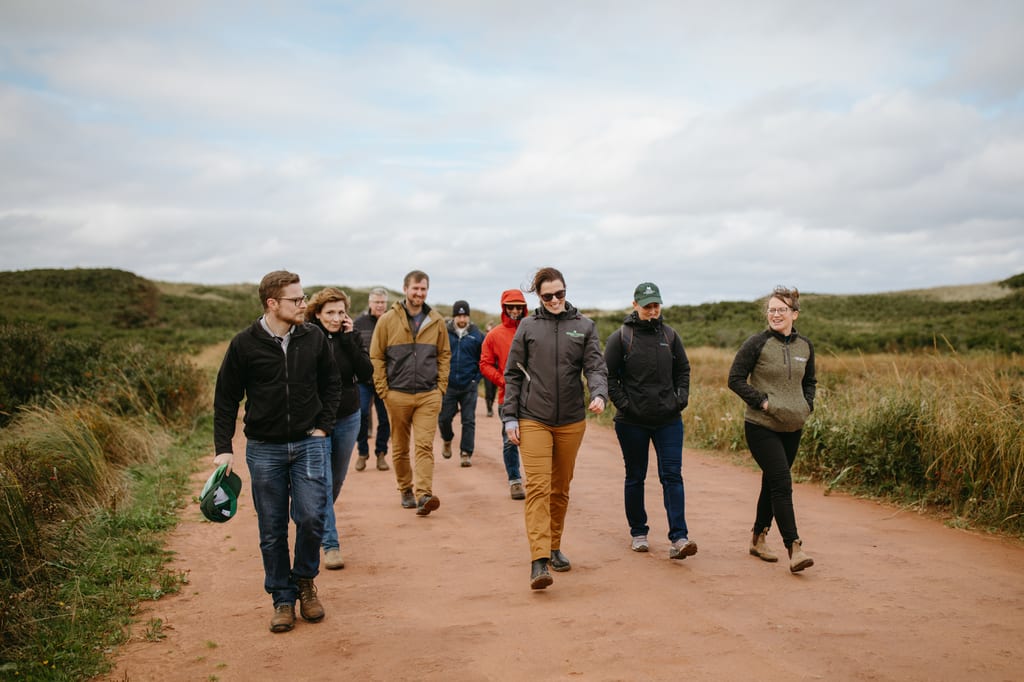 A group of people walking along a dirt path through a grassy landscape under a cloudy sky.