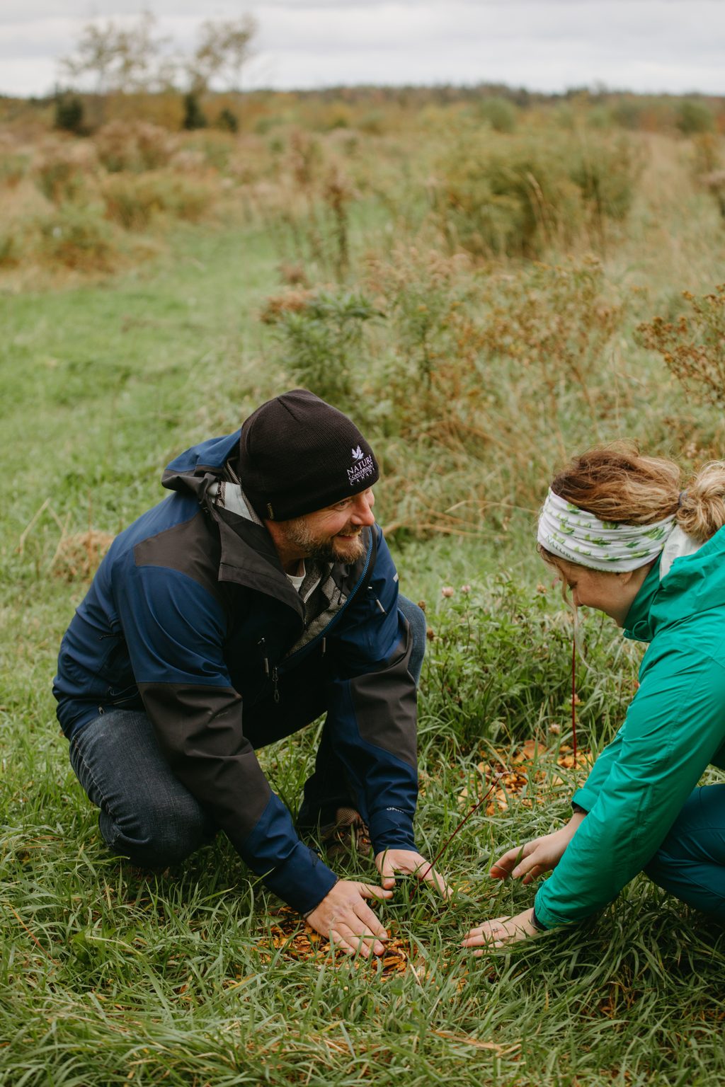 Two people kneel in a grassy field, examining the ground.