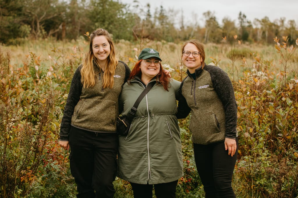 Three women in outdoor clothing pose for a photo in a field of tall grasses and shrubs.