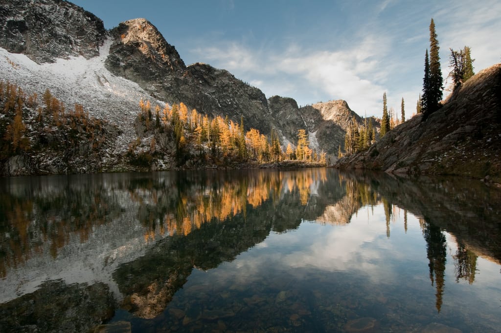 A mountain lake reflects the rocky peaks and golden larch trees in autumn.