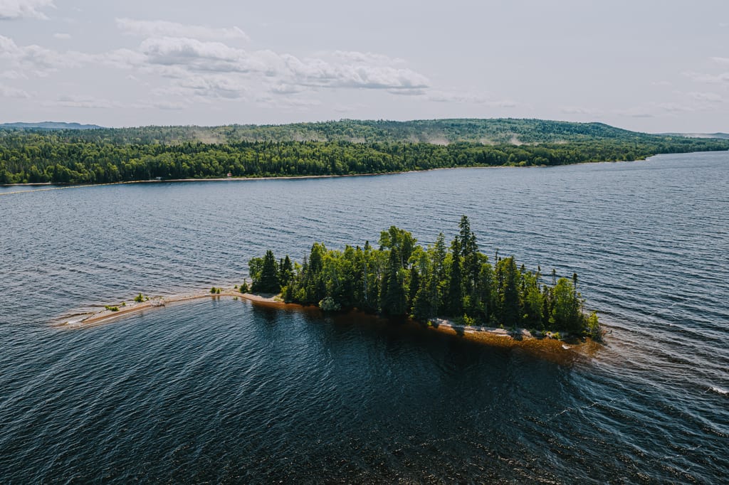 A small forested island sits in the middle of a large lake under a blue sky.