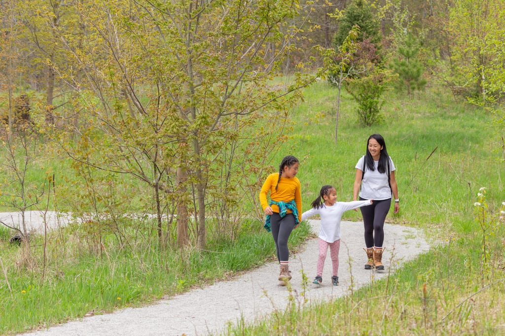 A woman and two children walk along a gravel path through a grassy, wooded area.