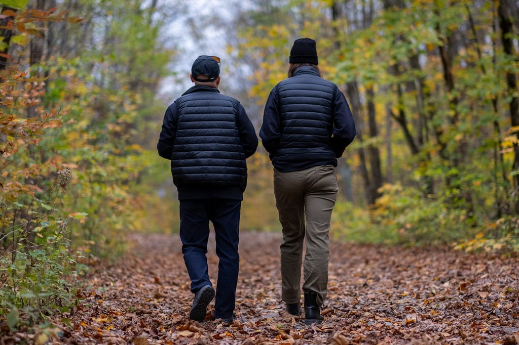 Two people walk along a leaf-covered path in a forest during autumn.