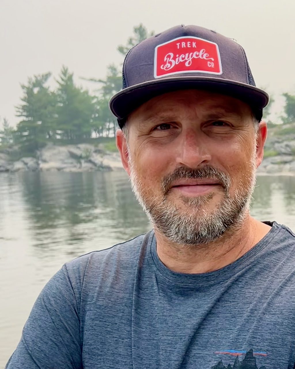 A man wearing a cap poses in front of a lake and a rocky shoreline.