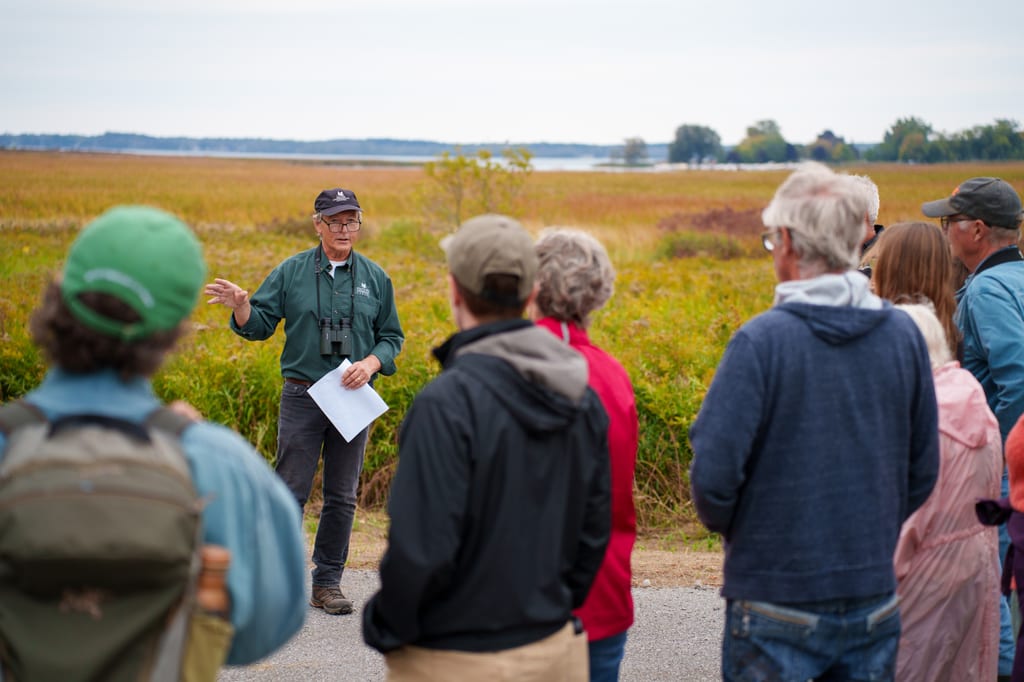 A guide speaks to a group of people in a grassy field near a body of water.