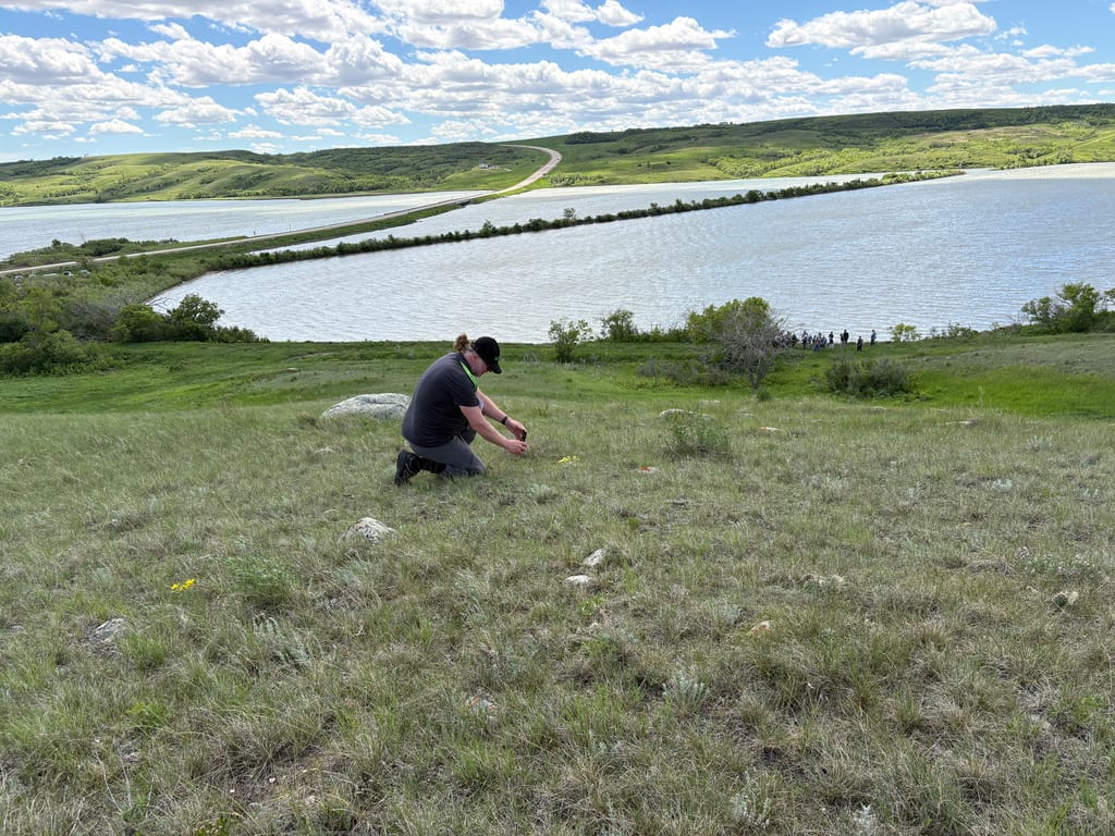 A person crouches in a grassy field overlooking a large lake and rolling hills.