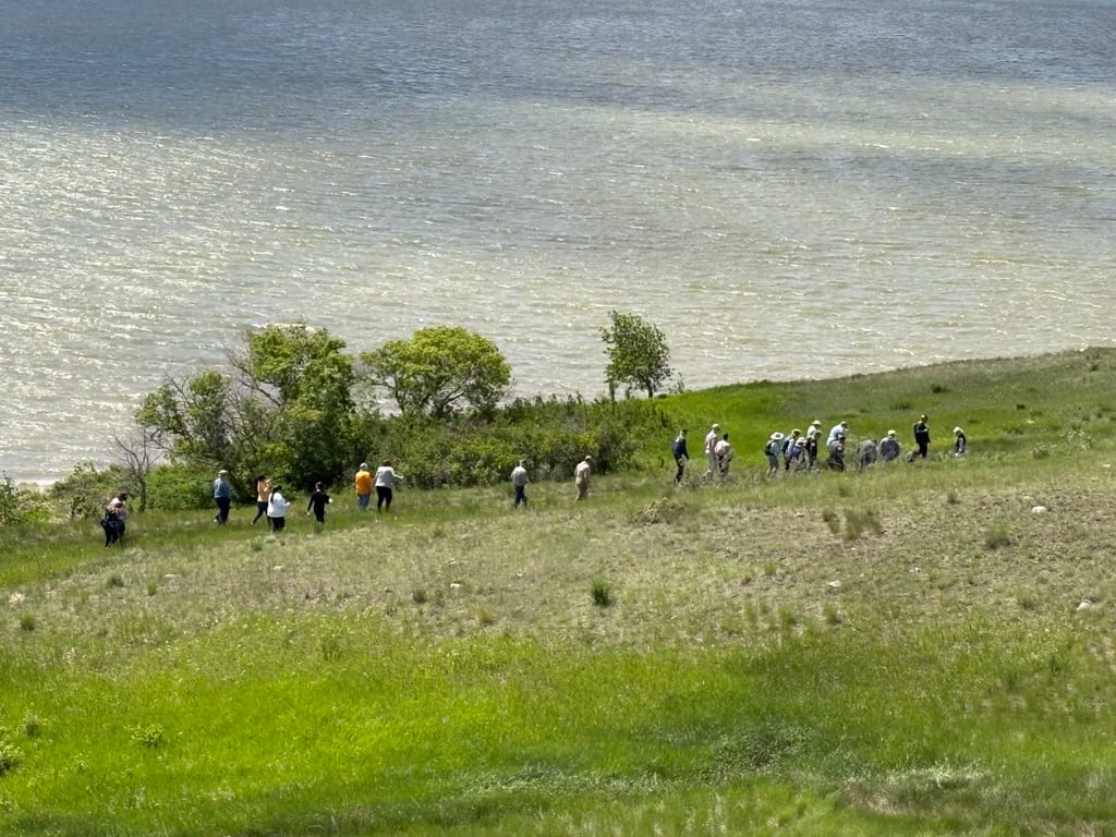 A group of people walks along a grassy hillside overlooking a large body of water.