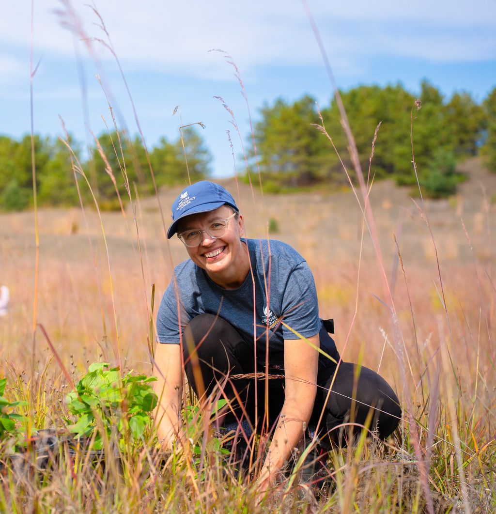 A person smiles while tending to small plants in a grassy field.