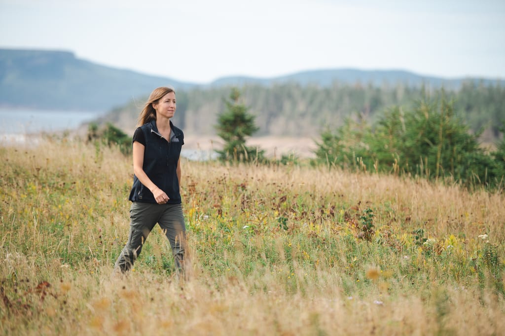 A NCC staff member walks through a grassy meadow with trees and hills in the background.