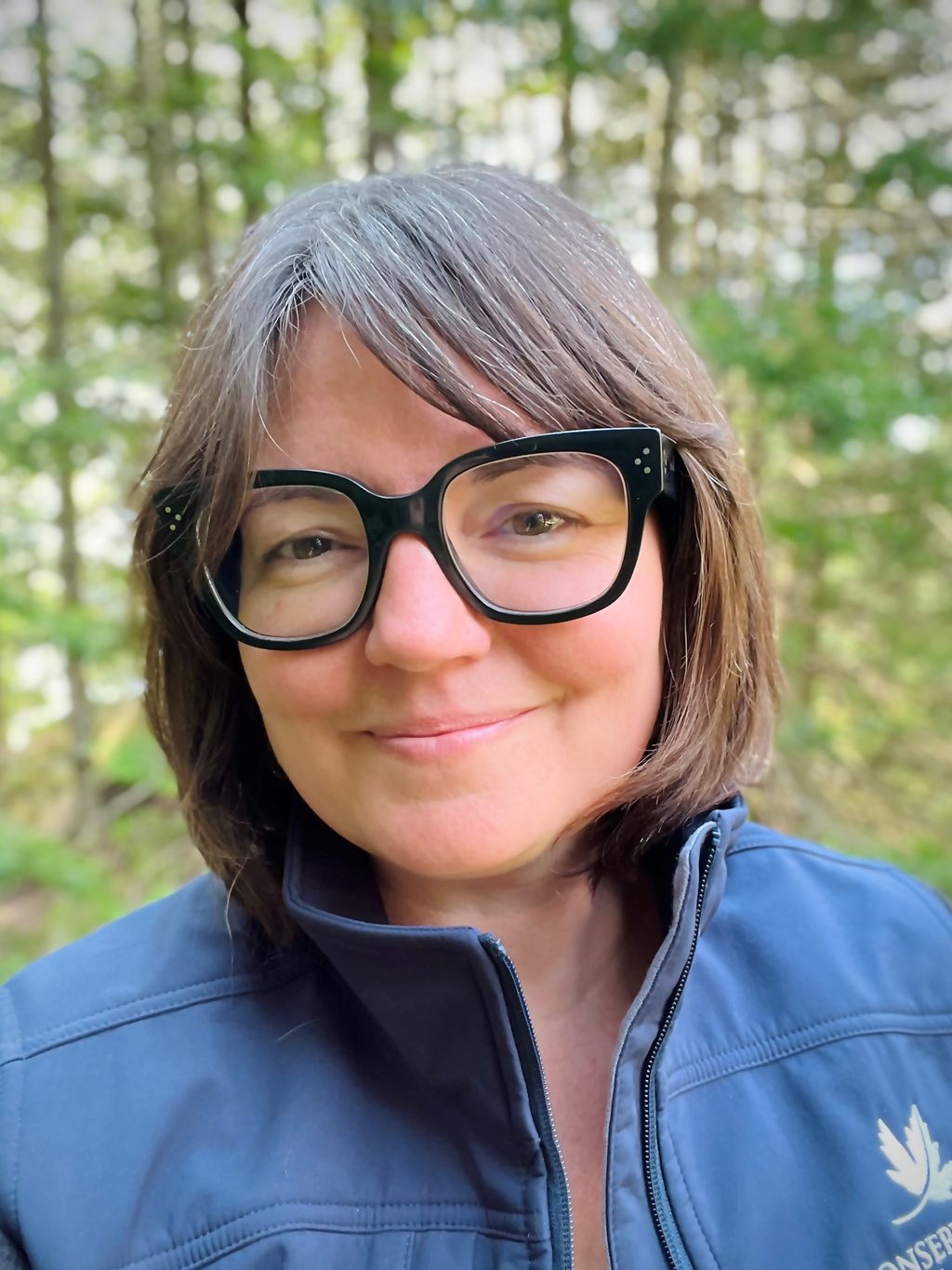 A woman wearing glasses and a blue jacket smiles in a forest.
