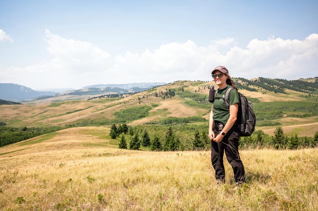 A woman with a backpack stands in a grassy meadow overlooking rolling hills and mountains.