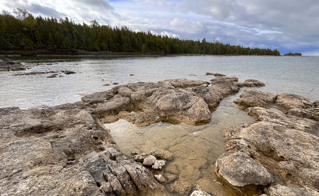 Rocky shores and shallow pools meet the calm water of a lake under a cloudy sky.