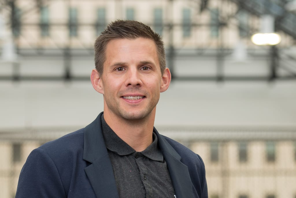 A man in a dark blazer and polo shirt poses for a portrait against a blurred background.