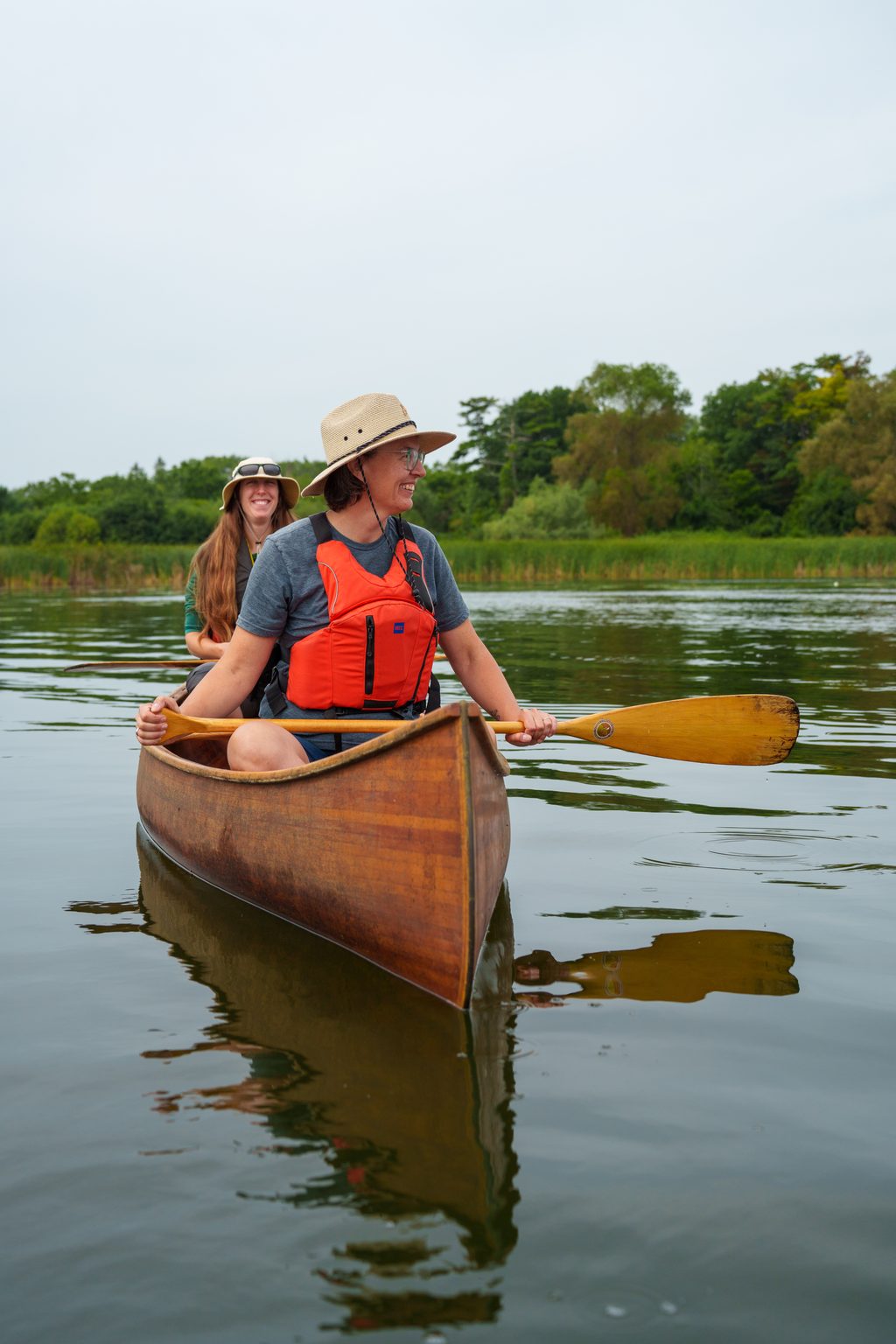 Two women paddle a wooden canoe on a calm lake surrounded by green trees.