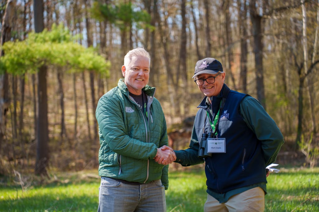 Two people shake hands in a sunny, wooded area.