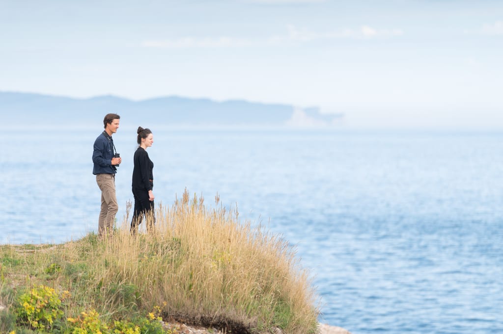 A man and a woman stand on a grassy cliff overlooking the ocean.