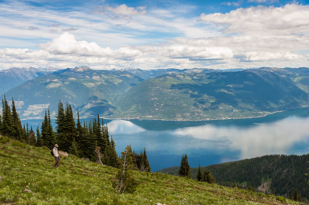 A hiker walks on a grassy mountain slope overlooking a large lake and distant mountains.