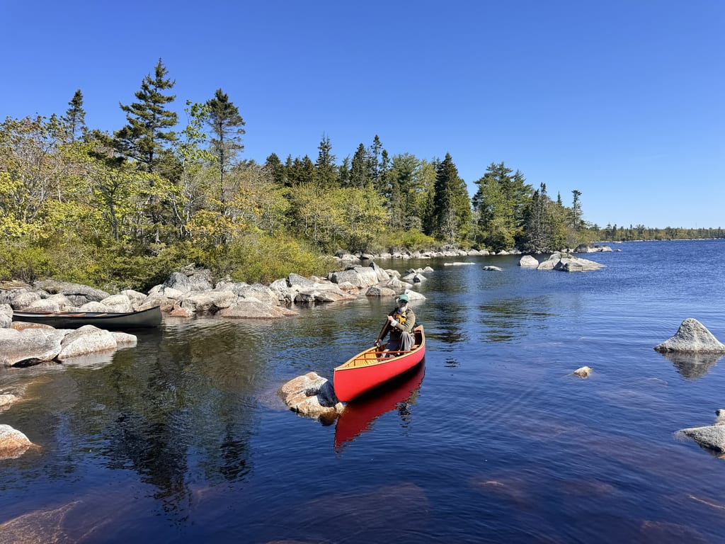 A person in a red canoe paddles on a clear lake surrounded by rocks and a forest.