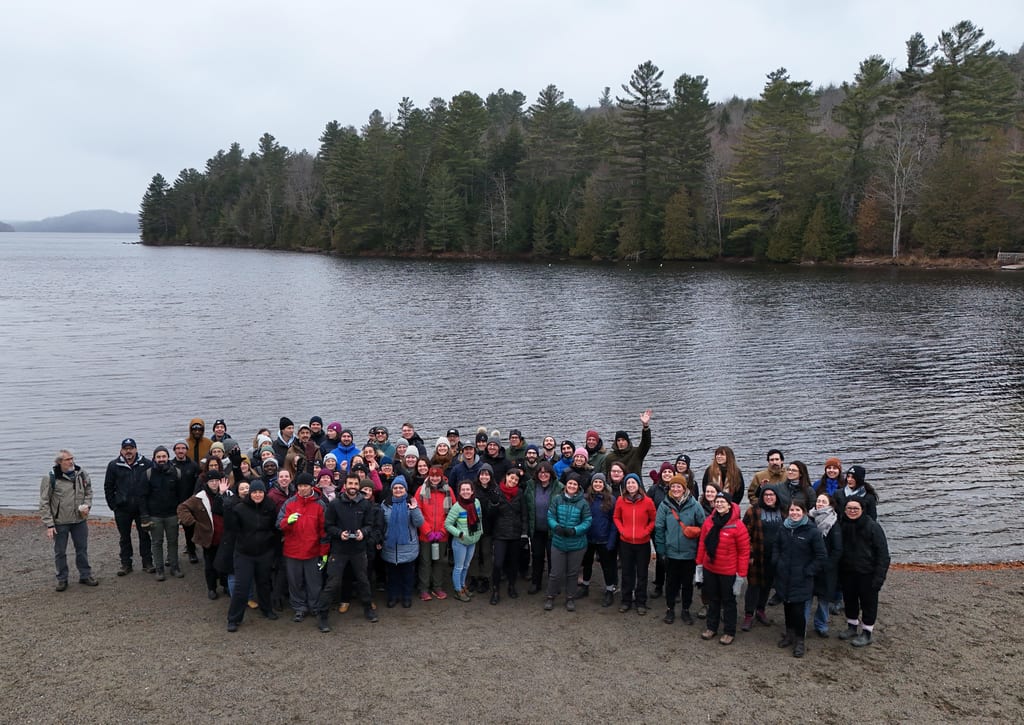 A large group of people posing for a photo on the shore of a lake with a forest in the background.