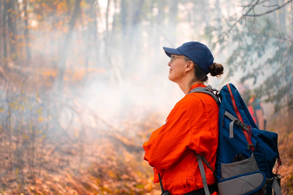 A person in an orange jacket and blue cap looks through the smoke in a forest during autumn.
