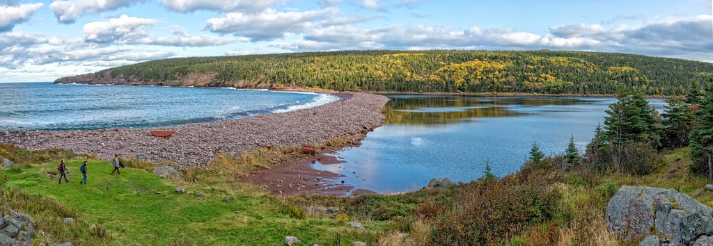 A person walk along a grassy slope overlooking a pebble beach and a calm inlet surrounded by autumn forests.