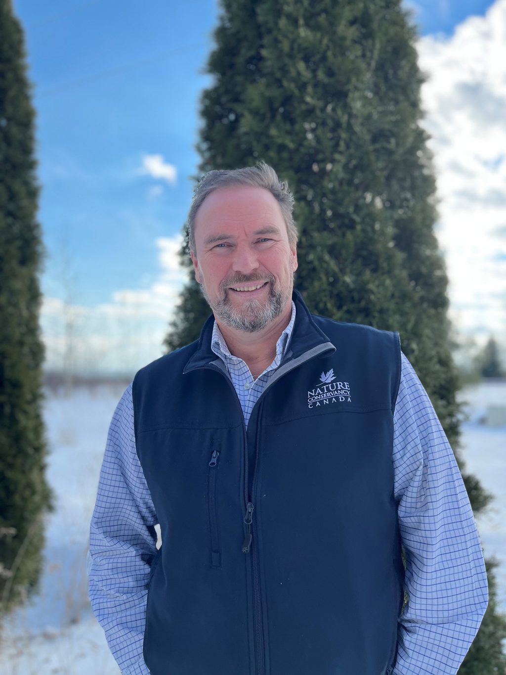 A man wearing a Nature Conservancy Canada vest stands in a snowy landscape with evergreen trees.