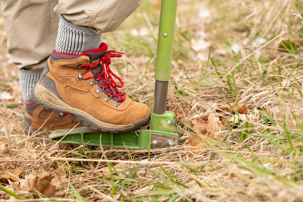A person wearing a hiking boot steps on a green tool in a field of dry grass and green shoots.