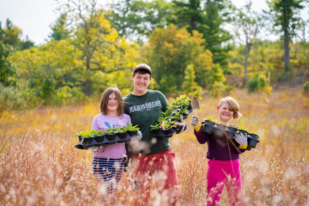 Three people hold trays of seedlings in a field with autumn foliage in the background.