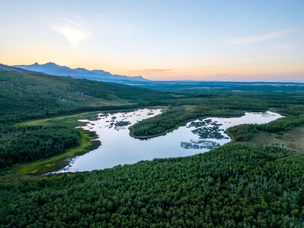 An aerial view of a lake surrounded by dense green forest and mountains under a sunset sky.