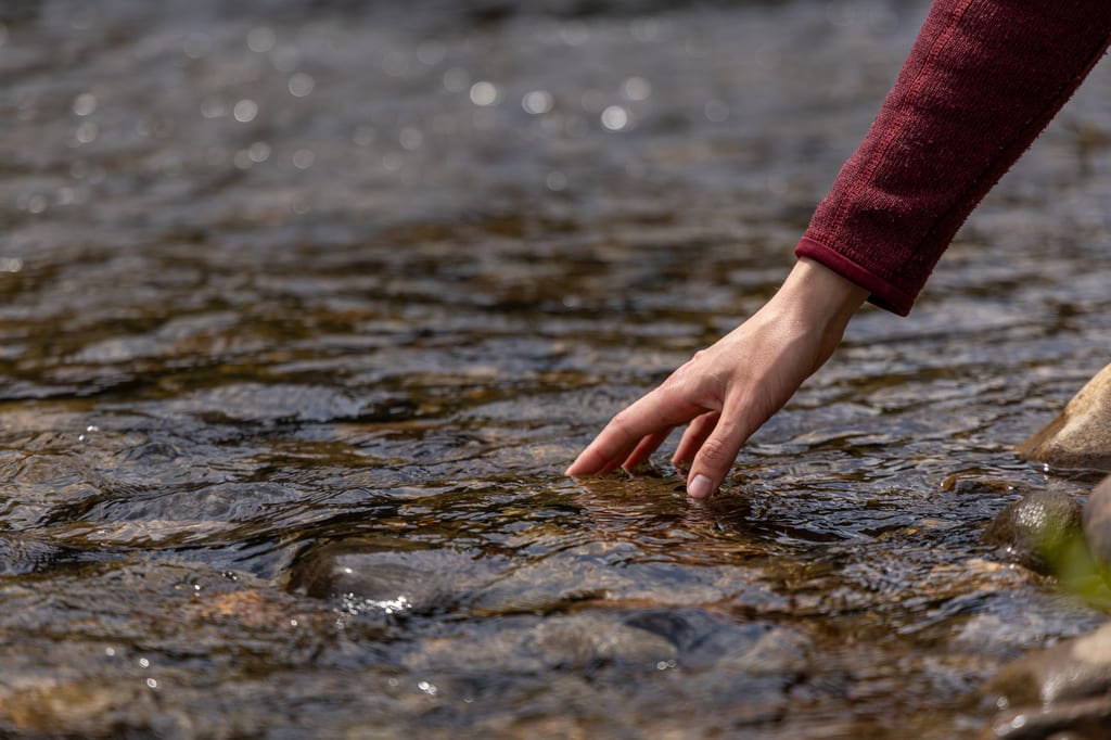 A hand reaches into the clear, shallow water of a rocky stream.