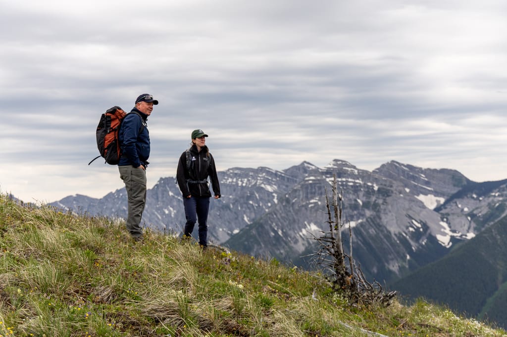 Two hikers walk along a grassy mountain slope with rocky peaks in the background.