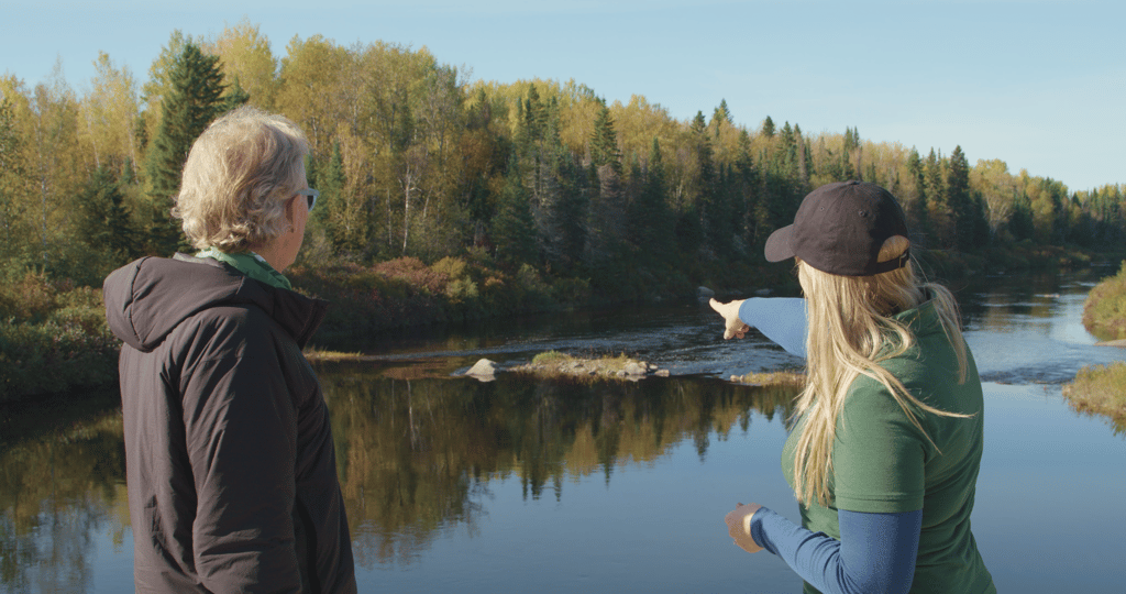 Two people stand by a river in a forest during autumn, with one person pointing across the water.
