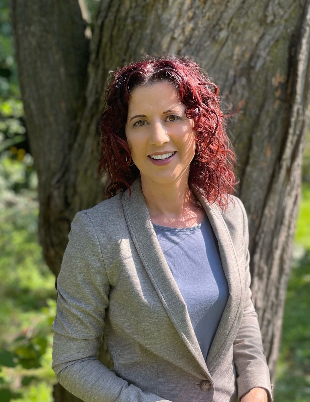 A woman with curly red hair poses in front of a large tree trunk.