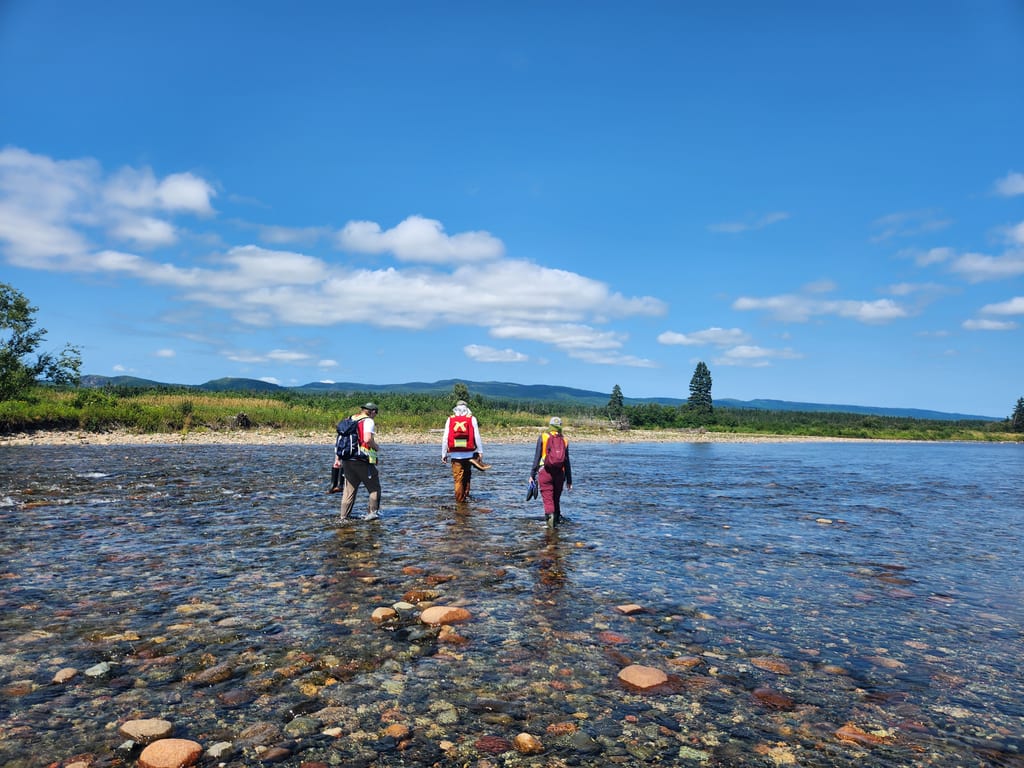 Trois personnes marchent dans une rivière peu profonde et rocailleuse sous un ciel bleu.