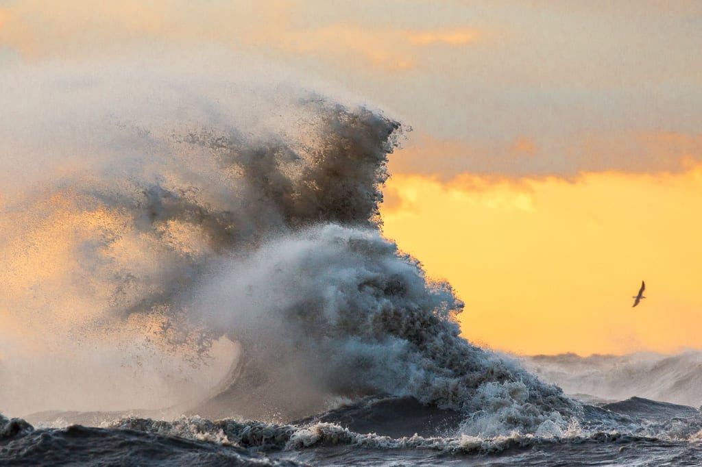 A large wave crashes in the ocean under a golden sunset sky.