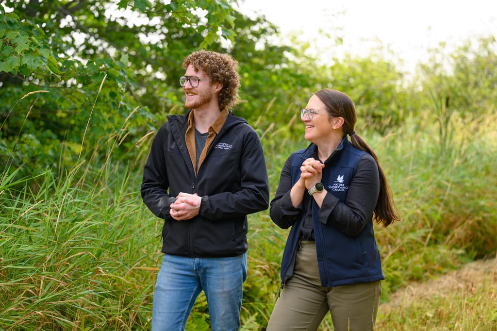 Two people stand in a field of tall grass and green foliage.