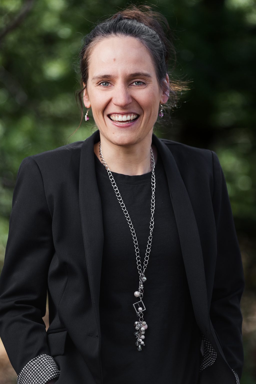 A smiling woman wearing a black blazer stands in front of green foliage.