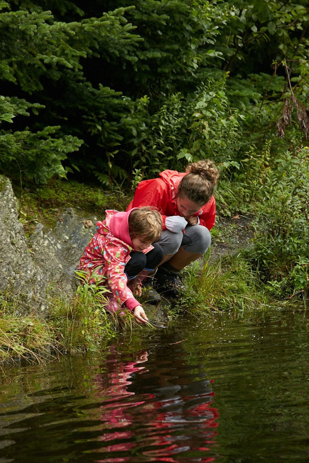 Two children in colorful jackets crouch by the edge of a pond in a lush green forest.