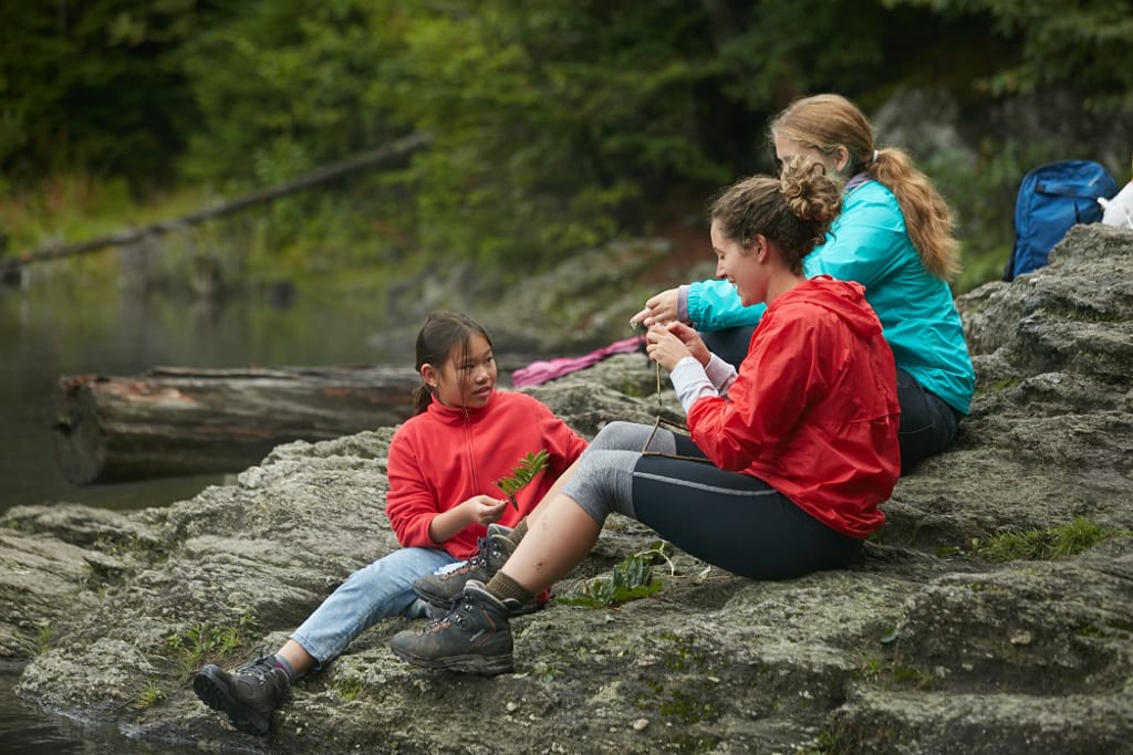 Three young women sit on rocks by a lake, examining a small plant.